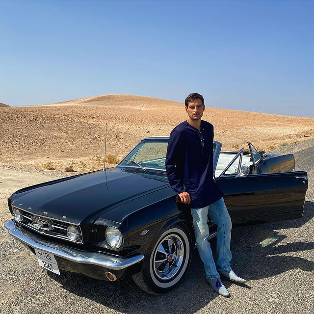 Brice Bexter in a dark blue vest and light blue jeans posing in front of his car.